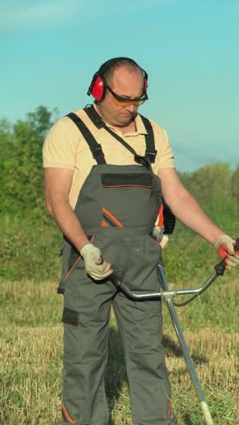 Landscaper in protective workwear looks down while holding and operating a gasoline powered brush cutter, moving through tall grass in an open field beneath a clear blue sky