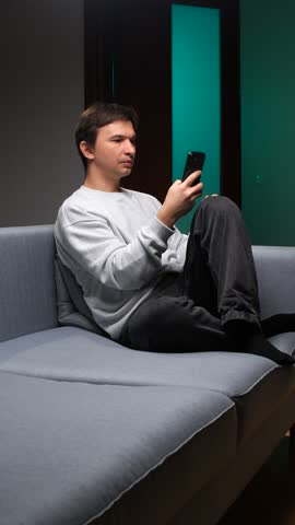 Vertical video man cheering with clenched fist while watching an online sports match on his smartphone, celebrating victory and success from the comfort of his home sofa