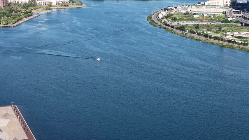 Aerial drone view of a white speedboat cruising on a vast blue lake in Zhengzhou, China. The boat leaves a long white wake on the calm water. Green shorelines and a modern city in the background under a sunny sky.