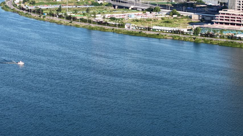 Aerial drone view of a white speedboat cruising on a vast blue lake in Zhengzhou, China. The boat leaves a long white wake on the calm water. Green shorelines and a modern city in the background under a sunny sky.