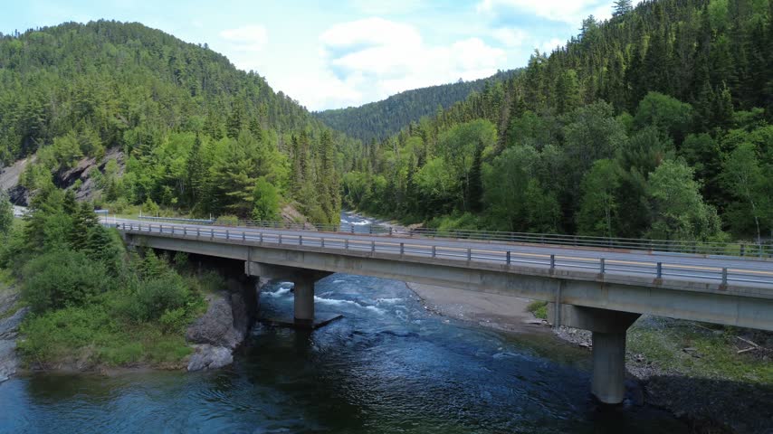 Cinematic drone view of a highway bridge spanning the Assemetquagan River, surrounded by forested slopes and scenic terrain in Quebec