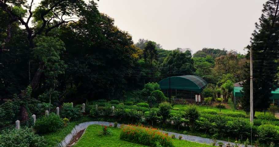 A scenic landscape view of a botanical garden featuring flower beds, a greenhouse, and dense trees under a soft, overcast sky.