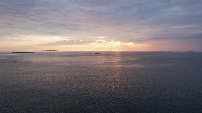 Aerial view of a peaceful coastal seascape at sunset, with warm sunlight reflecting across the water and subtle sun rays breaking through layers of dramatic clouds. Calm open water and distant islands create a tranquil atmospheric evening scene along the Norwegian coast.