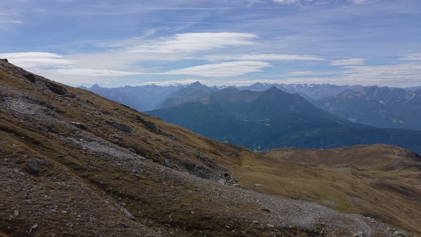 Drone reveal from rocky mountain grassland toward distant alpine peaks and a broad valley in Tyrol, Austria, beneath thin layered clouds.