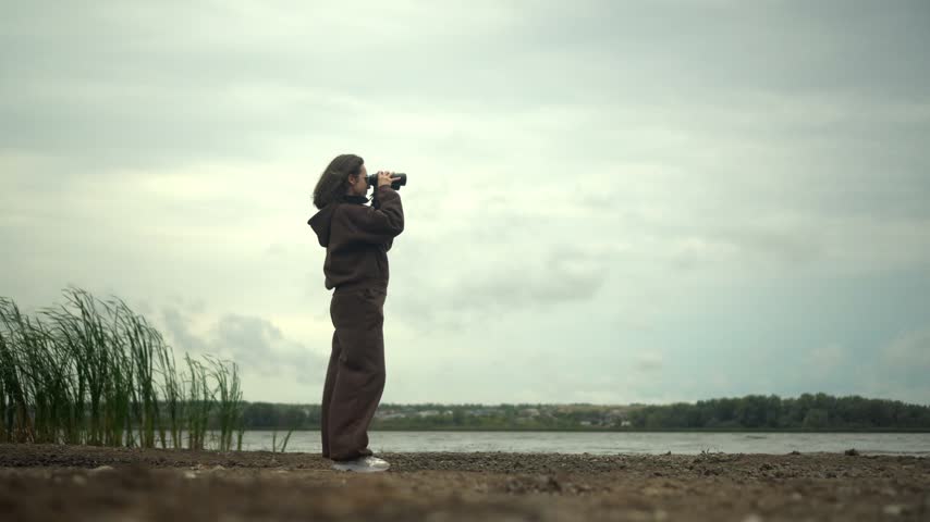 Young Woman in Glasses Looking Through Binoculars at the Opposite Shore of a Lake in Cloudy Weather, Wearing a Brown Tracksuit, Side View, Wide Shot