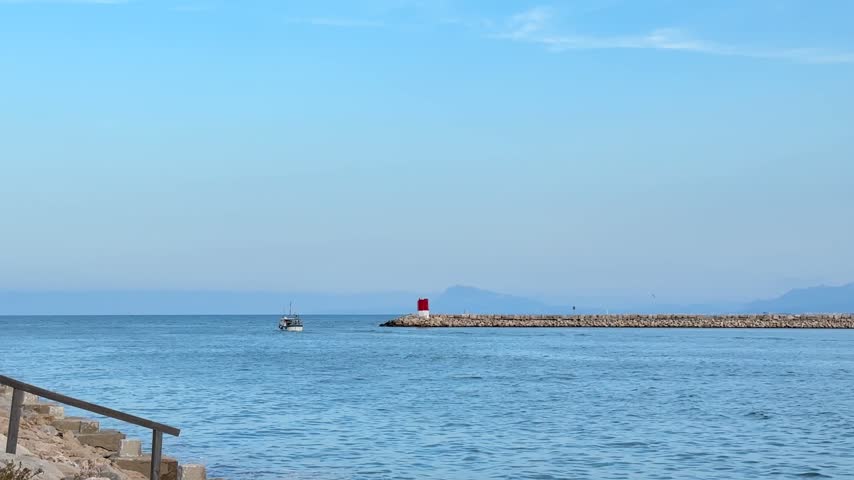 Flat calm Mediterranean water with a red navigation beacon at the end of the long breakwater and hazy mountains beyond in Gandia, Spain.
