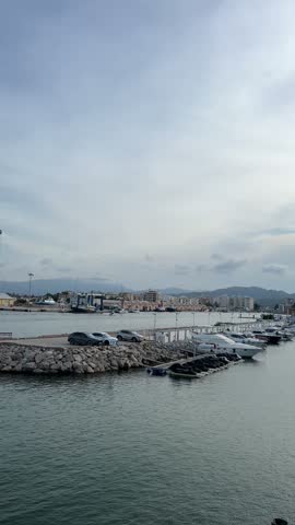 Slow vertical pan across the marina and promenade as the evening sun sinks behind clouds over the resort skyline in Gandia, Spain.
