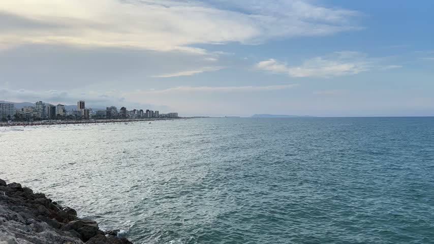 View across the Mediterranean towards the distant resort skyline of Gandia, framed by dark rocks in the foreground, Spain.