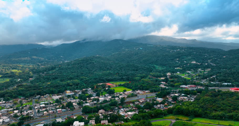 City lies at the foot of the verdant mountains. Dramatic cloudscape cover the sky over the green hills. Drone footage above Puerto Rico, USA on cloudy day.