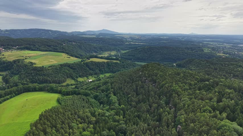 Broad aerial landscape view shows rolling hills, patchwork fields, dense forest edges, and distant mountain ridges beneath soft partly cloudy light.