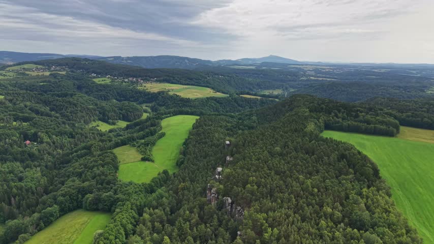 Broad aerial panorama shows rolling green hills with dense trees, patchwork meadows, and rocky outcrops. Fields and small settlements stretch toward hazy mountain ridges under soft overcast light.
