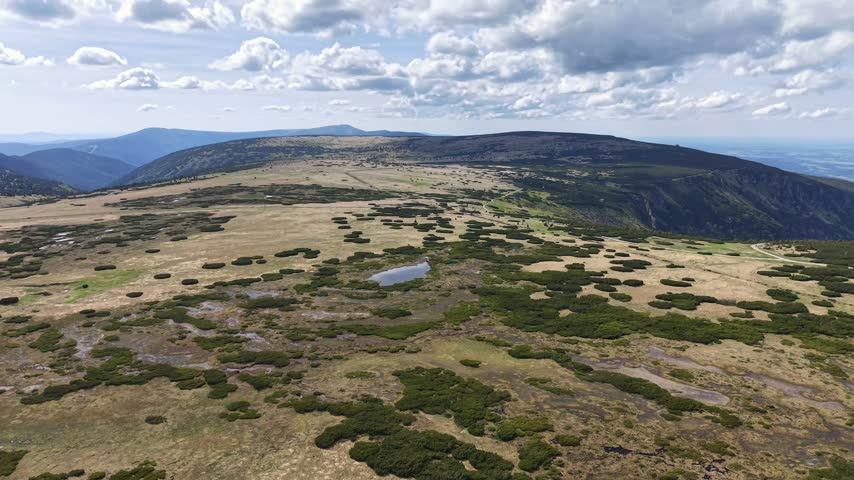 Wide aerial panorama of open mountain plateau with rounded ridges, scattered low shrubs, and reflective pools across brown and green grassland under partly cloudy skies.