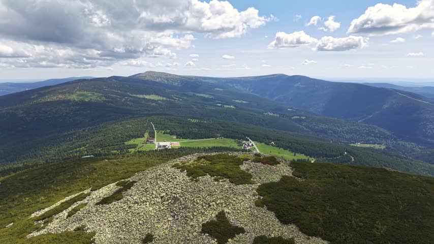 High ridge viewpoint shows rocky foreground, green patchwork fields, small clusters of buildings, winding roads through forested valley, layered mountains on horizon.