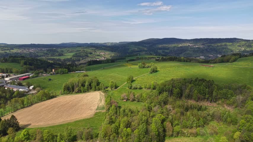 High angle panorama of rolling patchwork fields with hedgerows, scattered farms, winding rural roads, and distant village beneath clear blue daylight, lush greenery bright in sun.