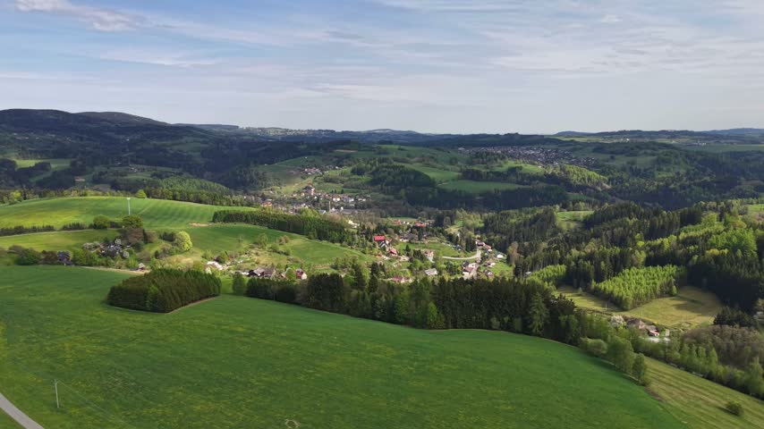 Wide aerial view shows rolling green hills, patchwork fields, small villages, and dense woodlands under bright lightly clouded sky.