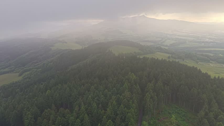 Aerial view shows dense conifer forest on ridge above rolling green hills and pastures, while village fades into mist in sunrise atmosphere. Warm light highlights terrain and distant horizon.