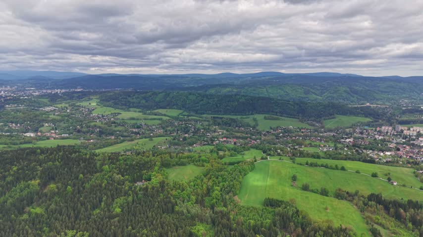 Sweeping overhead view shows quilted fields, forested slopes, and scattered buildings along winding roads, bordered by meadows and tree lines under stratified moody clouds.