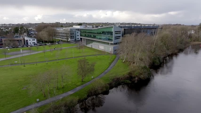An aerial drone shot showcasing the modern sustainable engineering building at the University of Galway, Ireland, situated along the scenic River Corrib with green campus grounds.