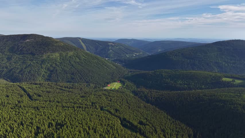 Elevated aerial view of meadow valley with red roof cottages, dense evergreen forest, and layered ridges. Bright sunlight and soft shadows create depth beneath clear blue sky for outdoor themes.