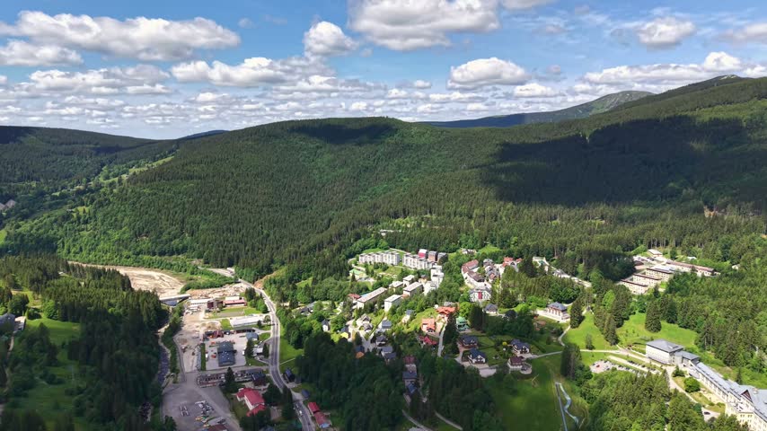 Elevated aerial scene shows mountain town bordered by dense pine forest and rolling hills. Homes and rooftops sit along valley edge near winding roads, green lawns, and tree canopy under bright sky.