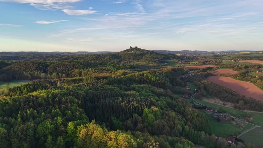 Aerial perspective shows ancient ruins on hill crest above patchwork woodland and farmland, with meandering roads and small settlements under bright blue sky and warm sunlight.