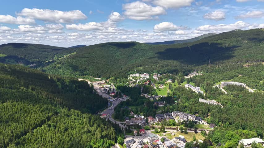 Sweeping aerial panorama shows tranquil valley town nestled among dense pine forests and tall mountain ridges, with winding streets, rooftops.