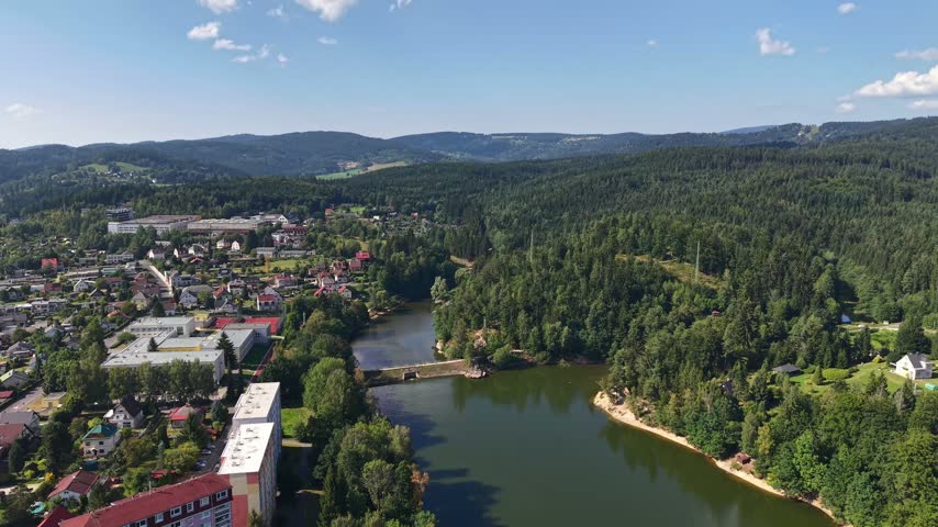 Clear daylight aerial perspective shows riverside town beside winding river, red roofs clustered near bridges, surrounded by pine forest, green hills, and distant ridges under clear blue sky.