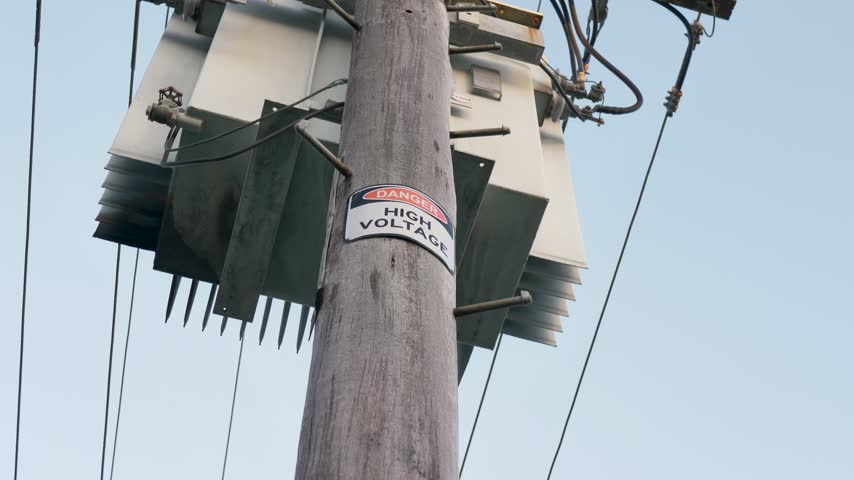 Slow motion landscape of a pole-mounted electrical distribution transformer attached to a wooden utility pole with power lines and a high voltage danger warning metal sign on a suburban town street