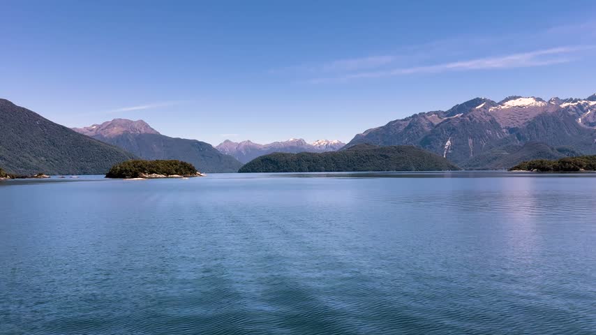 Small forested islets break the surface of Lake Manapouri while steep bush clad ridges and a distant snow capped range close the horizon, Fiordland. Wide shot
