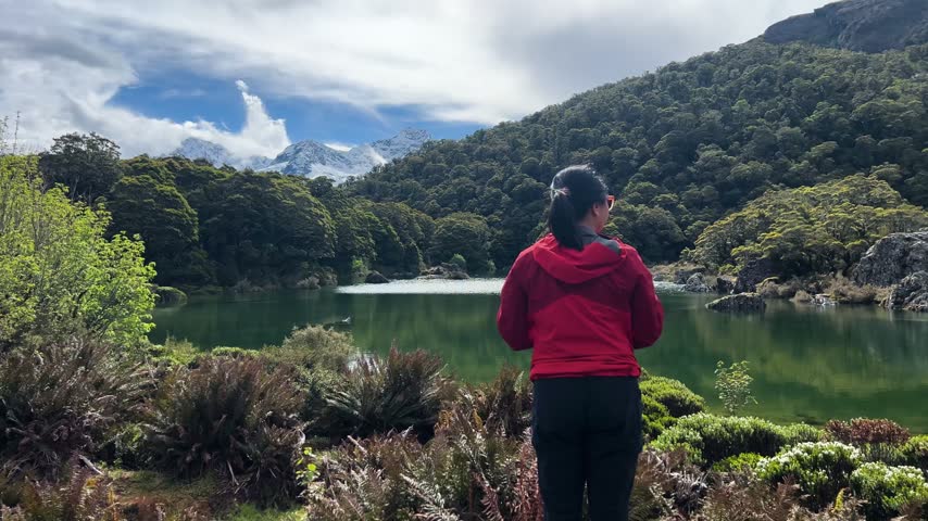 A walker in a red jacket stands at the edge of Lake Mackenzie on the Routeburn Track, New Zealand, green water and beech forest running to a snow capped peak beyond. Wide shot