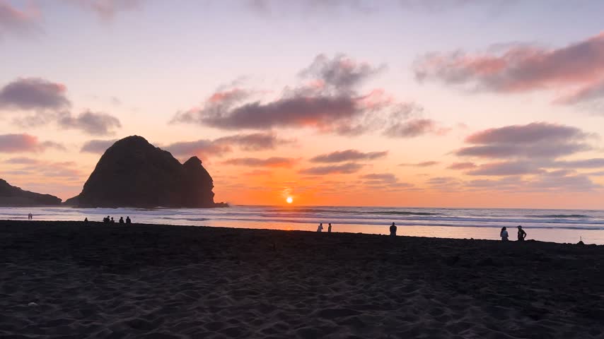 The sun drops to the horizon beyond Lion Rock at Piha, New Zealand, walkers small on the black sand and pink cloud spread across the evening sky. Wide shot