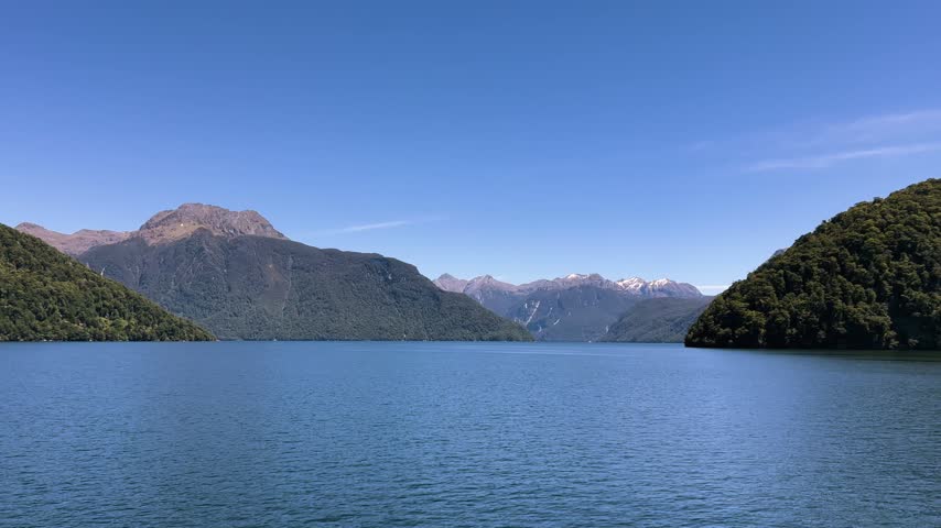 Wide basin of Lake Manapouri with a pointed forested peninsula on the right, brown tussock tops on the left and a snowy range far up the lake. Wide shot