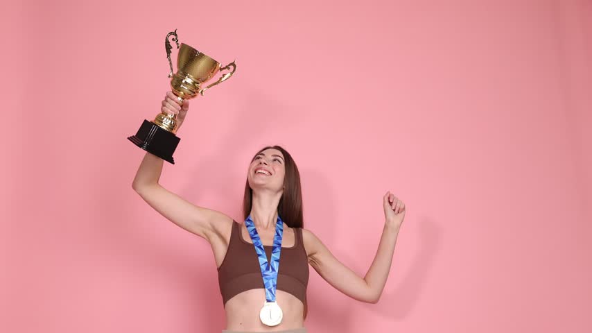 Happy woman with gold medal and trophy cup on pink background, low angle view. Slow motion effect