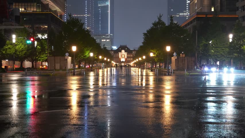 Rainy night view of Tokyo Station in Tokyo, Japan, featuring illuminated buildings, pedestrians with umbrellas and colorful city lights reflecting on the wet pavement.