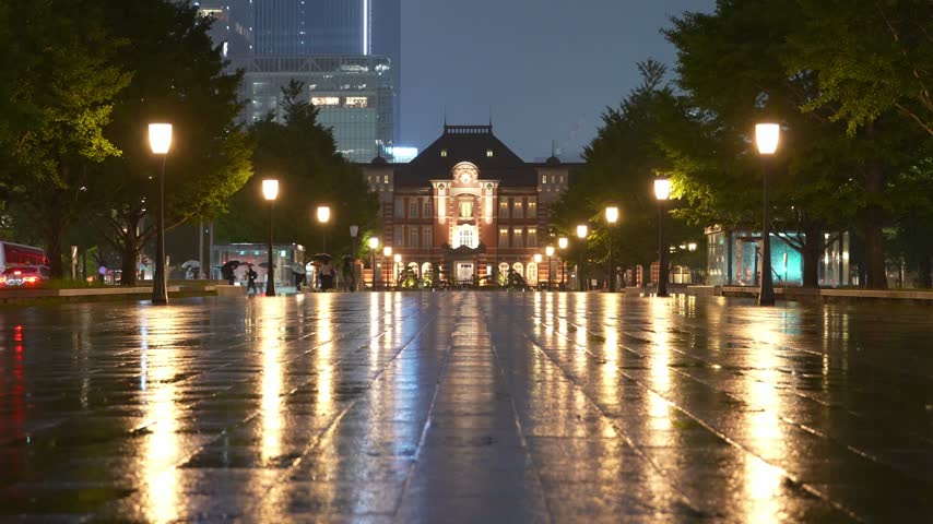 Rainy night view of Tokyo Station in Tokyo, Japan, featuring illuminated buildings, pedestrians with umbrellas and colorful city lights reflecting on the wet pavement.