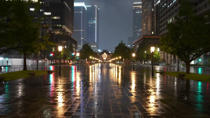 Rainy night view of Tokyo Station in Tokyo, Japan, featuring illuminated buildings, pedestrians with umbrellas and colorful city lights reflecting on the wet pavement.