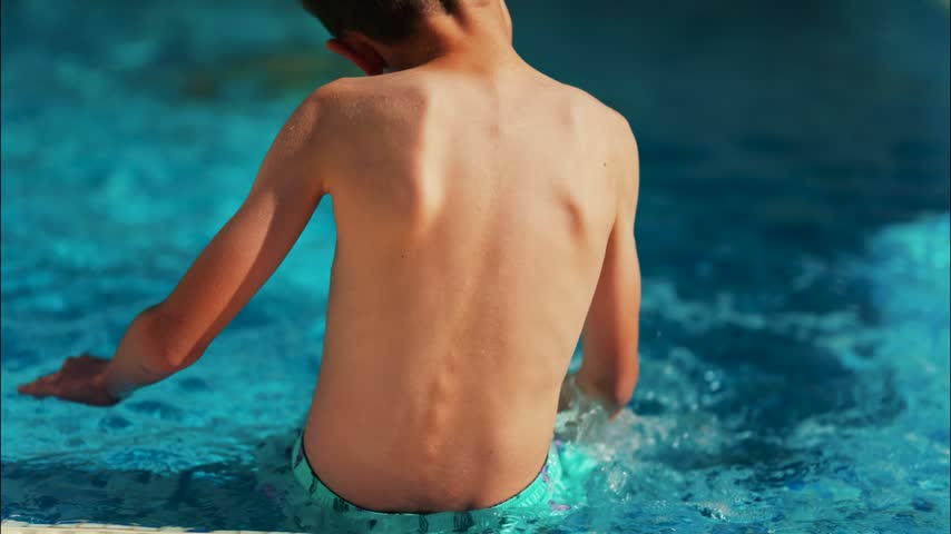 Child playing in swimming pool water with hands creating splashes during sunny summer day outdoor leisure activity.