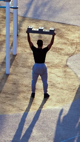 Athletic man performing overhead weightlifting exercises using heavy construction block in outdoor playground area during golden hour.