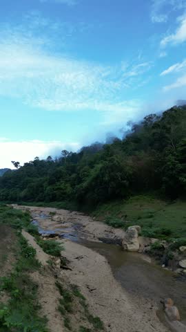 A scenic landscape featuring a partially dry sandy riverbed with a shallow stream, flowing alongside a dense green forested mountain covered in morning mist under a blue sky.