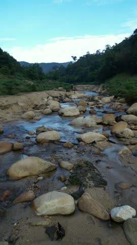 A scenic landscape featuring a partially dry sandy riverbed with a shallow stream, flowing alongside a dense green forested mountain covered in morning mist under a blue sky.