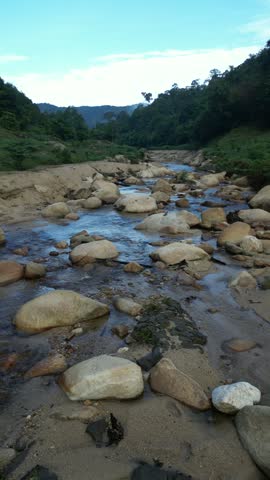 A scenic landscape featuring a partially dry sandy riverbed with a shallow stream, flowing alongside a dense green forested mountain covered in morning mist under a blue sky.