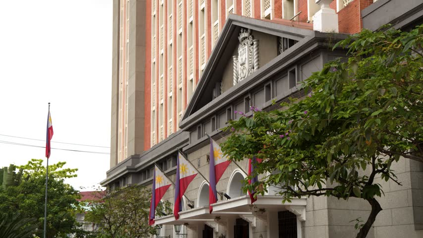 Slow motion footage capturing the exterior of a stately administrative building in Manila adorned with multiple Philippine flags waving gently against a cloudy sky during daytime.