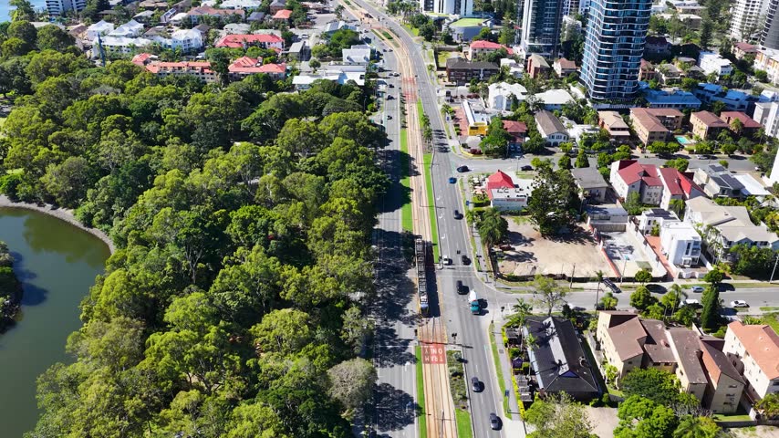 Aerial drone shot follows modern tram traveling along dedicated tracks through sunny coastal suburb. Lush green parkland lines one side while city buildings and urban architecture rise beyond.