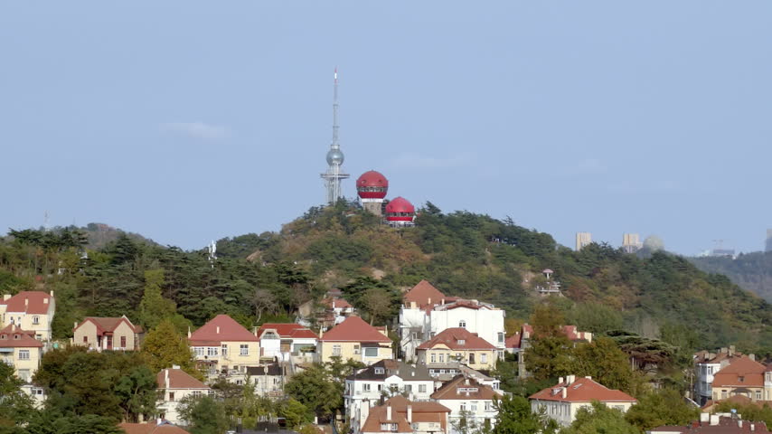 A hilly landscape features residential buildings and a tower with red domes under a clear blue sky.