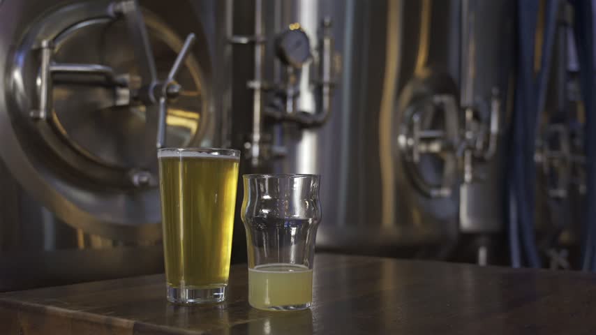 Close up shot of golden lager and cloudy wheat beer sitting on rustic table inside modern brewery with metal fermenters.