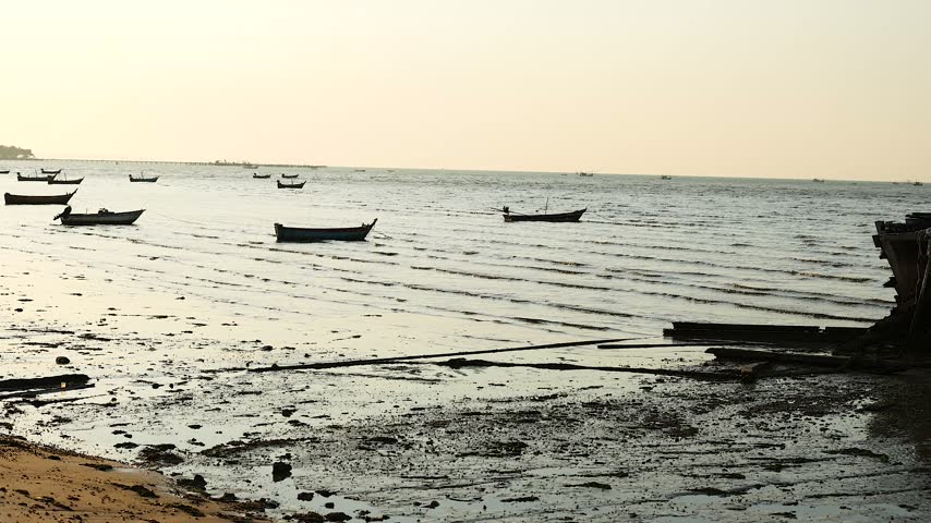 Small traditional fishing boats anchored in calm shallow coastal waters during sunset. Scenic landscape shot featuring gentle sea waves and muddy shoreline in peaceful golden hour light.