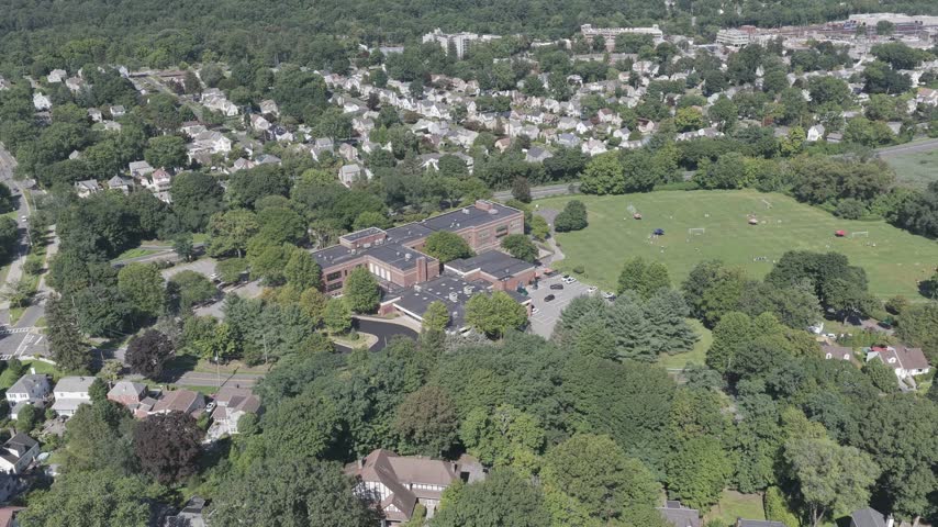 A steady drone view overlooks brick buildings beside a wide green playing field in White Plains, New York, framed by leafy trees, detached houses and residential streets in bright summer daylight.