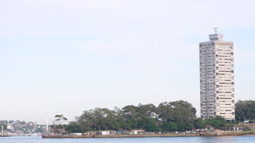 Tall residential high-rise rises above trees along the shoreline of Sydney Harbour. Sydney, New South Wales, Australia.