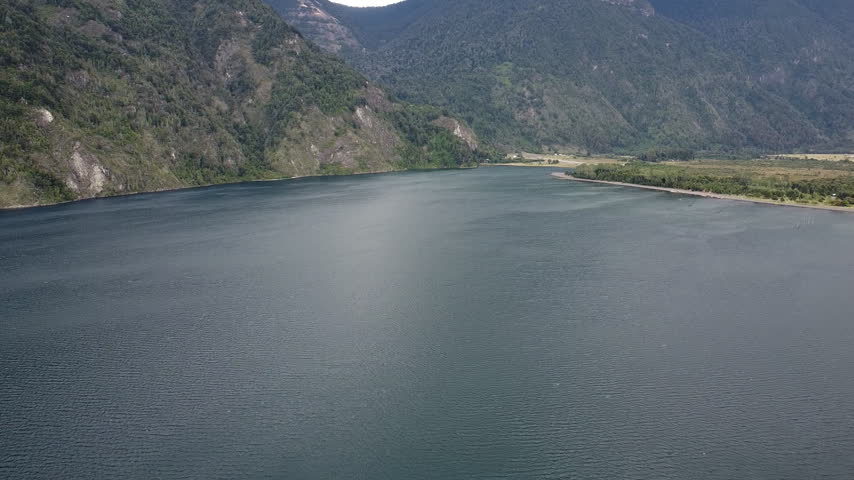Aerial wide view across a calm lake bordered by steep forested mountains, a narrow shoreline, and a green valley.