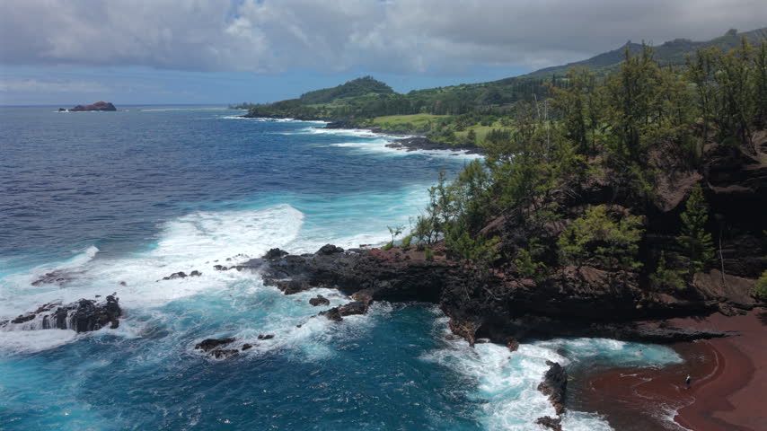 Aerial view of Kaihalulu Beach on Maui in the Hawaii island chain showing red sand cove sheltered by volcanic lava rock cliffs turquoise waves crashing and lush green hills above the ocean.